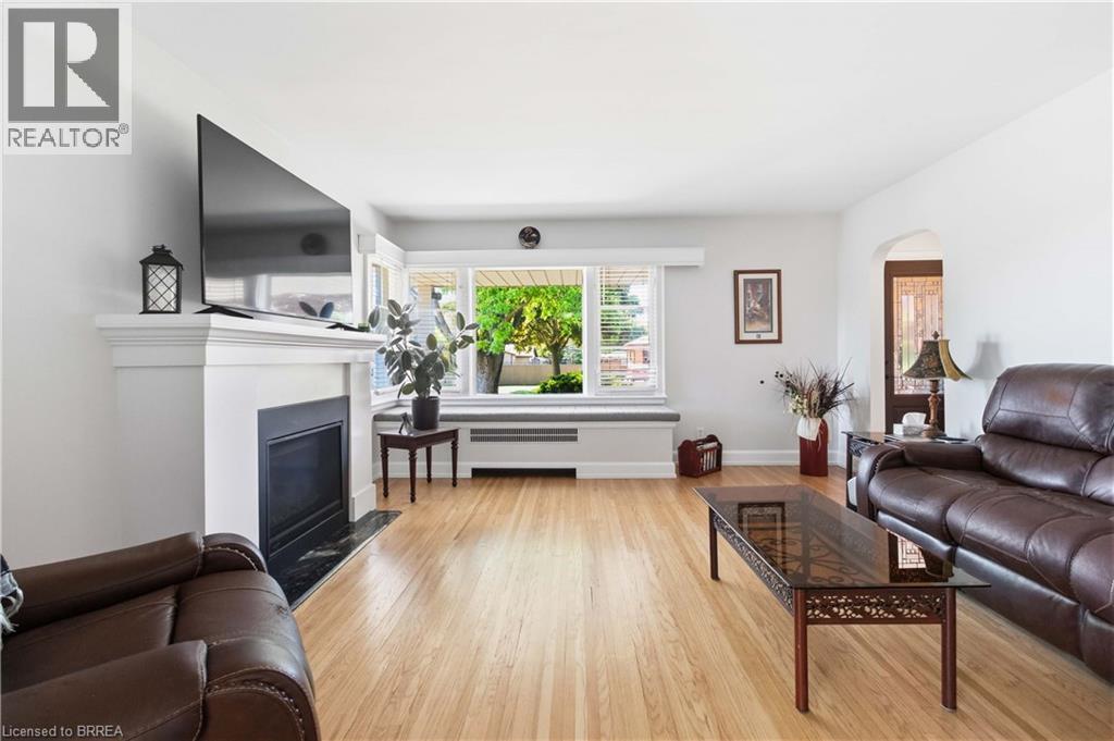 Bright living room featuring wood-finish flooring, a white mantel fireplace with a black surround, and a large multi-pane window with blinds - 134 Queensway Drive, Brantford, ON