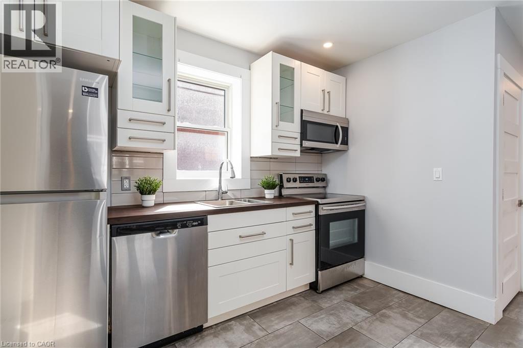 Compact kitchen featuring white cabinetry, stainless steel appliances, a tile backsplash, dark countertops, and gray tile flooring - 40 Woodland Avenue, Kitchener, ON