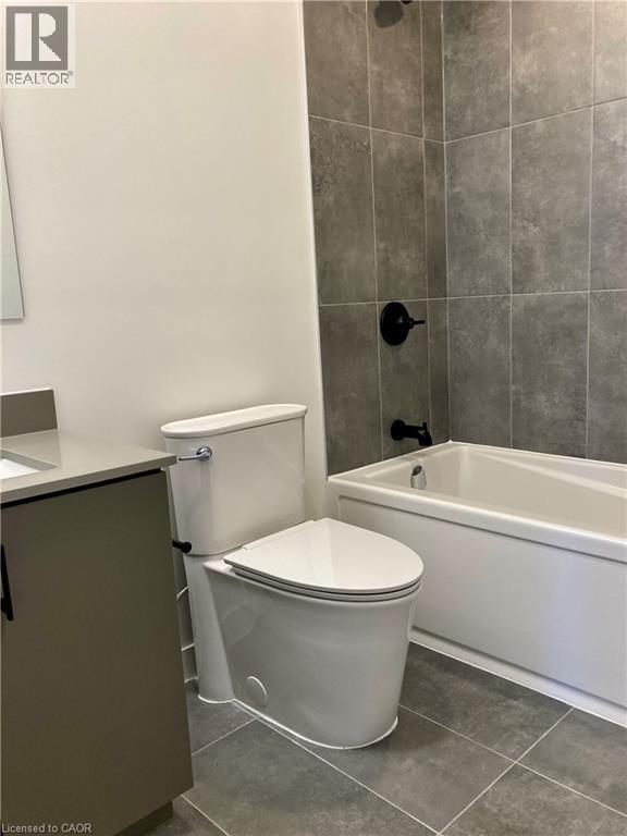 Bathroom featuring a white toilet, coordinating vanity with a light countertop, and a built-in bathtub with dark-toned wall tiling - 4878 Powers Common Unit# C-1105, Burlington, ON