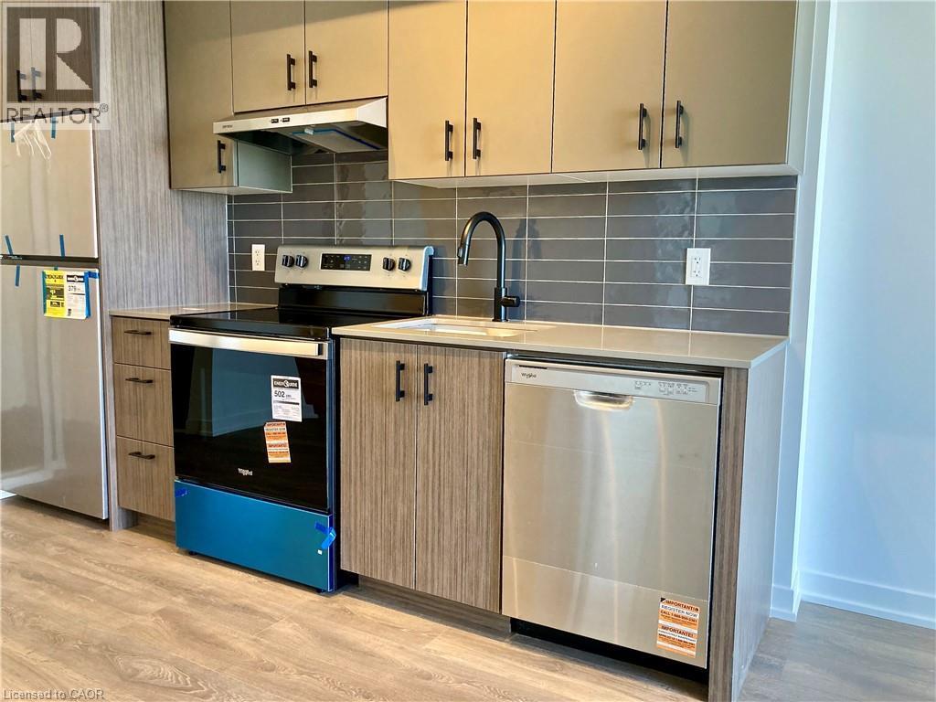 Modern kitchen featuring stainless steel appliances, grey subway tile backsplash, light-toned countertops, wood-finish flooring, and two-tone cabinetry with matte black hardware - 4878 Powers Common Unit# C-1105, Burlington, ON