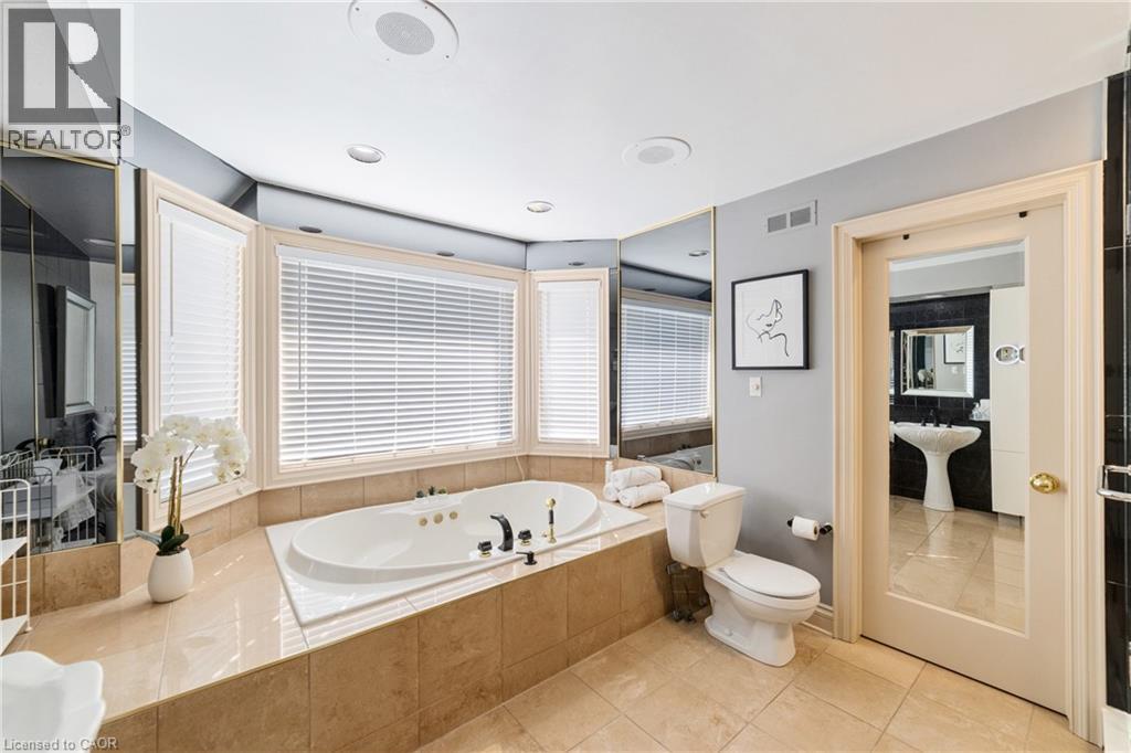 Bathroom featuring a built-in soaking tub with a tiled surround, bay windows with horizontal blinds, neutral floor tiling, and wall-mounted mirrors - 7775 St. Augustine Crescent, Niagara Falls, ON