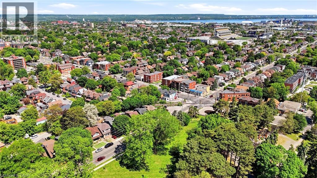 Expansive aerial view showcasing a verdant neighborhood with a mix of residential structures and multi-story buildings, abundant mature trees, a visible body of water in the distance, and an athletic - 968 Main Street E, Hamilton, ON