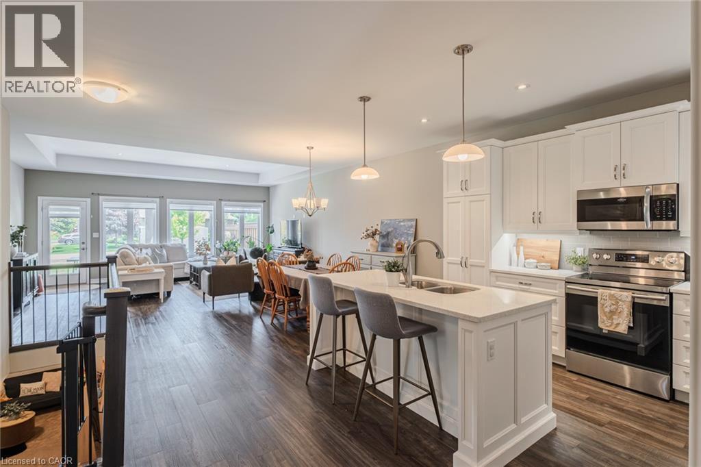Open-concept living space featuring wood-finish flooring, a contemporary kitchen island with seating, white shaker cabinetry, stainless steel appliances, and abundant natural light from multiple windo - 15 Winterberry Lane, Waterford, ON