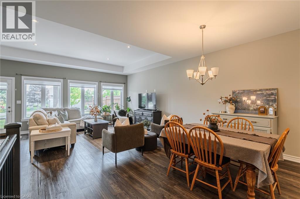 Open-concept living and dining areas featuring wood-finish flooring, a tray ceiling with recessed lighting, multiple large windows, and a classic chandelier over the dining space - 15 Winterberry Lane, Waterford, ON