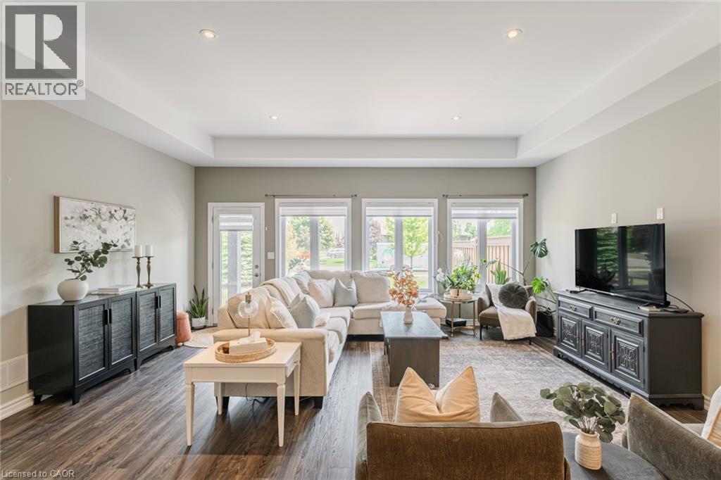 Spacious living area featuring wood-finish flooring, a tray ceiling with recessed lighting, and a wall of windows and an exterior door providing ample natural light - 15 Winterberry Lane, Waterford, ON
