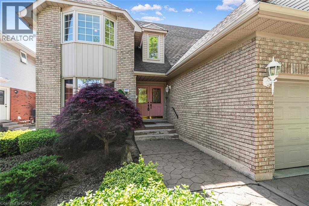 Brick exterior with a prominent bay window feature, dual entry doors, an attached garage, patterned concrete walkway, and established landscaping - 85 Citation Crescent, Ancaster, ON