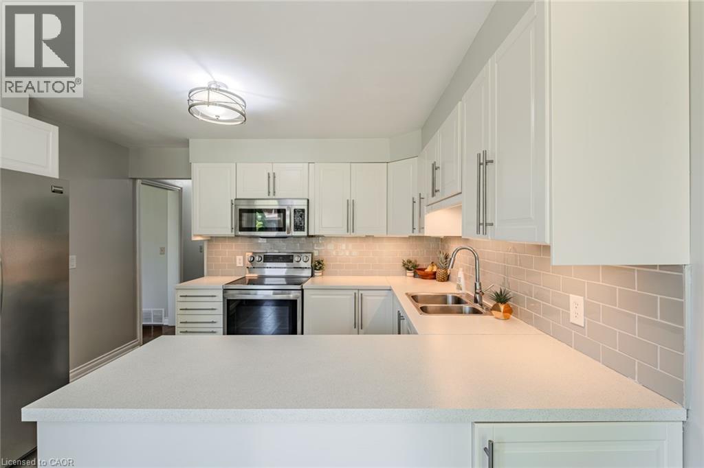 U-shaped kitchen featuring white cabinetry with brushed nickel hardware, light-toned countertops, a stainless steel double-basin sink with a gooseneck faucet, a subway tile backsplash, and stainless s - 141 Lynndale Road, Simcoe, ON