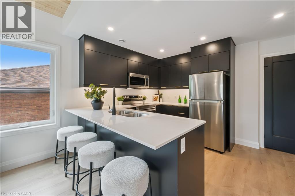 Contemporary kitchen with a central island featuring a white countertop and seating space, dark flat-panel cabinetry, stainless steel appliances, and light wood-finish flooring - 60 Arlington Boulevard Unit# 3, Kitchener, ON