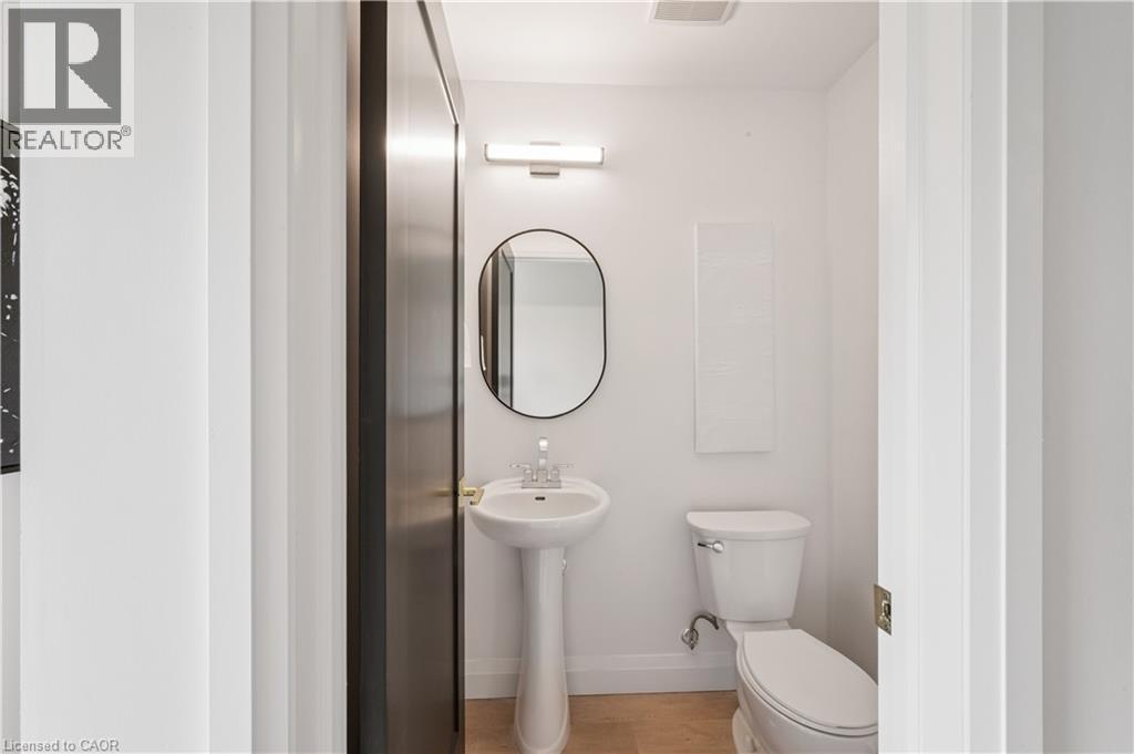 White-walled powder room featuring a pedestal sink with chrome faucet, a wall-mounted oval mirror, and a white toilet - 60 Arlington Boulevard Unit# 3, Kitchener, ON