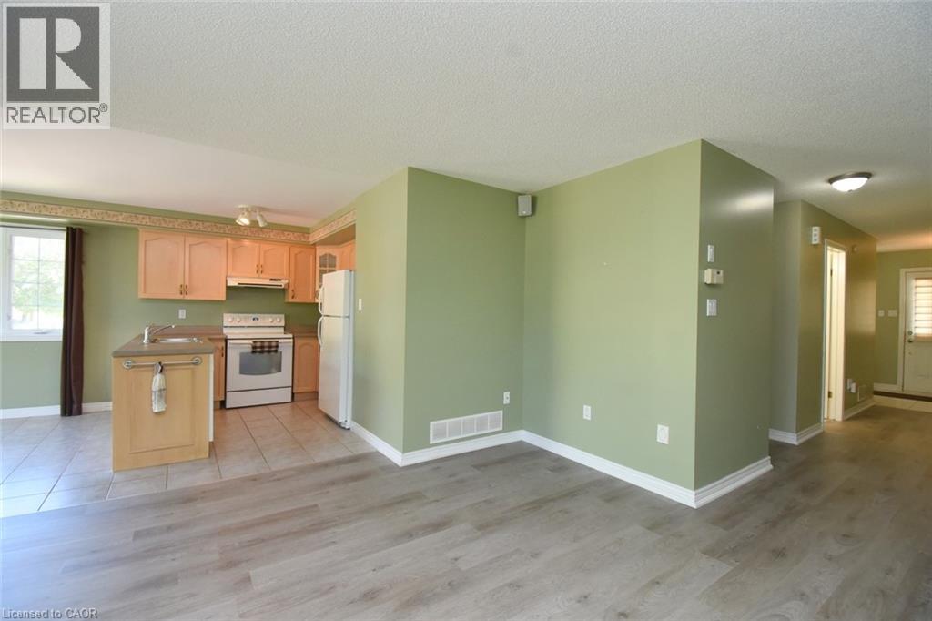 Open-concept kitchen and living area featuring wood-finish flooring, light wood cabinetry, a central kitchen island with a sink, white appliances, and a window providing natural light - 2 Harbour Drive Unit# 17, Stoney Creek, ON