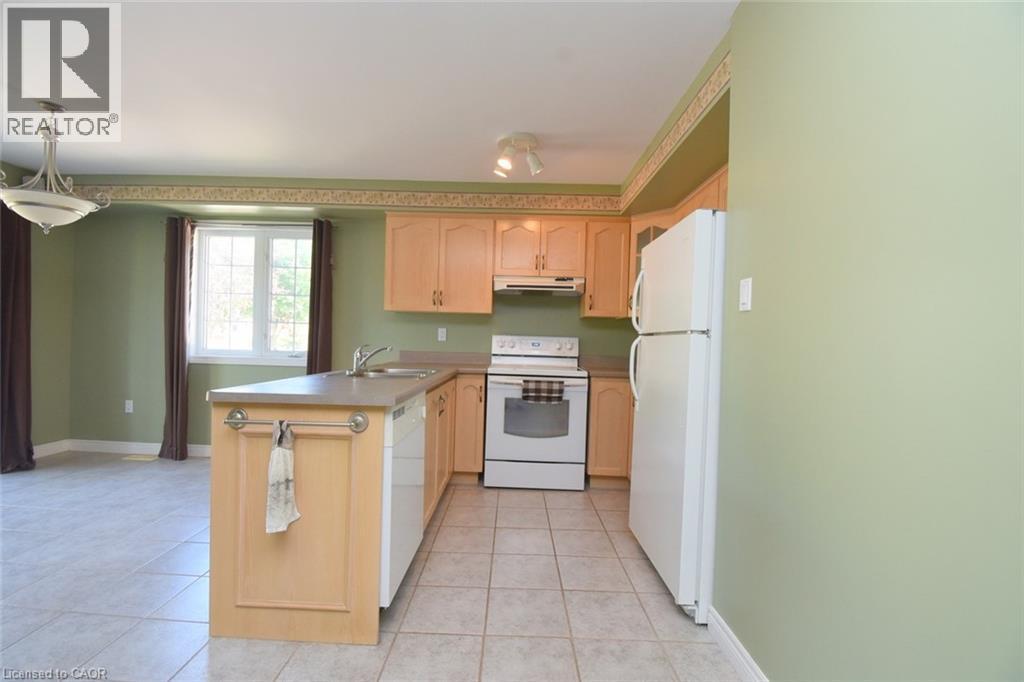 Kitchen featuring light wood-finish cabinetry, a central island with integrated sink, white appliances, and tiled flooring - 2 Harbour Drive Unit# 17, Stoney Creek, ON