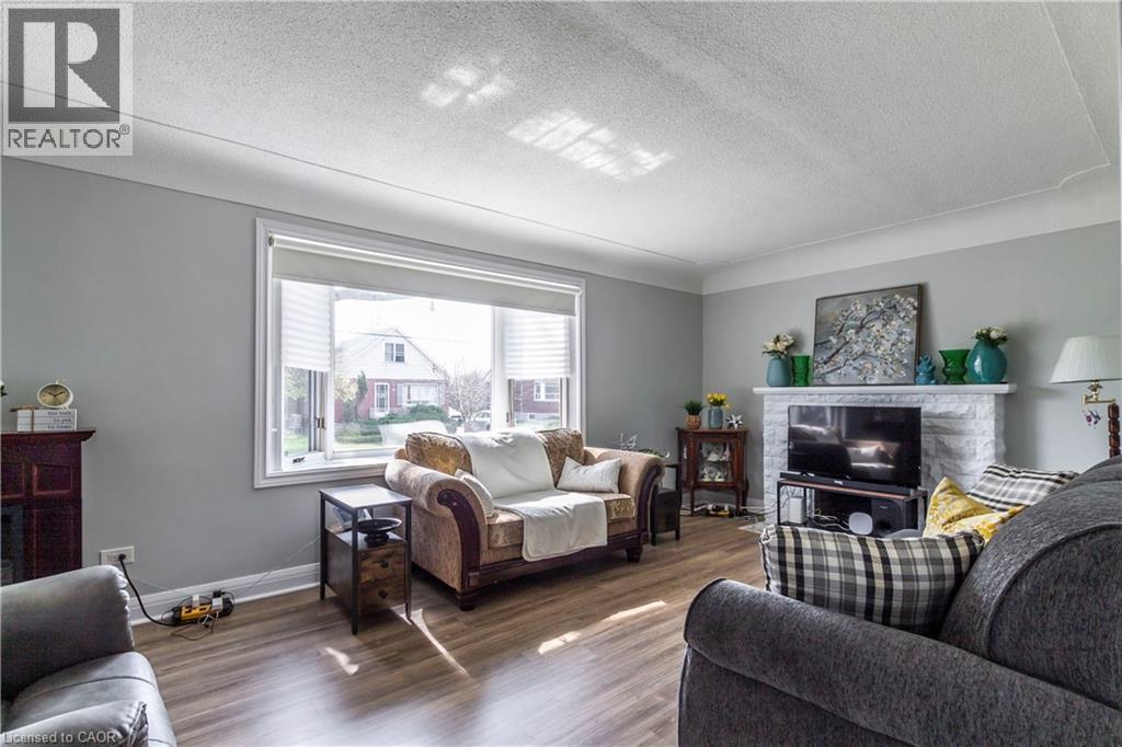 Spacious living area featuring light gray wall paint, crown molding, a large picture window with blinds, wood-finish flooring, and a stone-faced fireplace with a mantel - 20 West 1St Street, Hamilton, ON