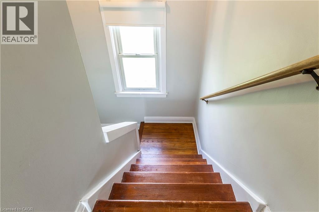 Staircase featuring wood-finish treads, a wood handrail with dark metal brackets, light neutral wall paint, white trim, and a double-hung window with white framing - 20 West 1St Street, Hamilton, ON