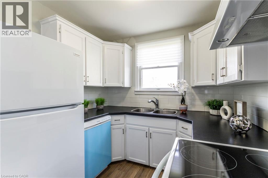 Modern kitchen featuring white cabinetry, dark countertops, a stainless steel sink, a tile backsplash, and wood-finish flooring - 20 West 1St Street, Hamilton, ON