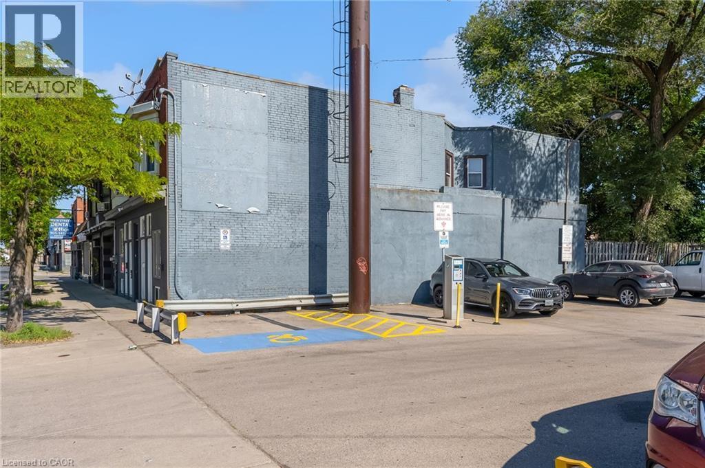 Street-level building featuring a brick exterior with boarded-up windows, a corner storefront with multiple window displays, and a paved parking area with designated accessibility spaces - 1371 Main Street E, Hamilton, ON - Outdoor