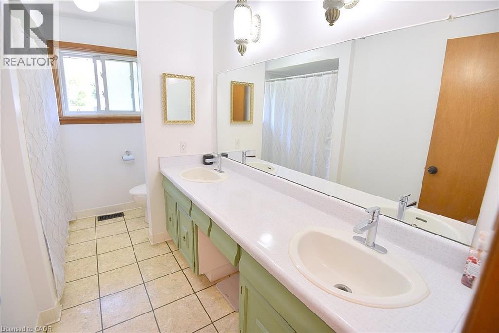Double vanity featuring dual undermount sinks with chrome faucets, a long wall-mounted mirror, and a wood-frame window - 372 Fennell Avenue W, Hamilton, ON