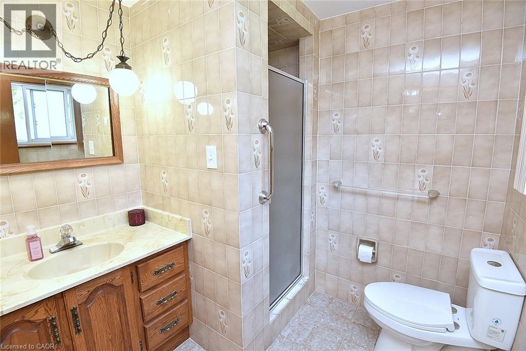 Bathroom featuring a walk-in shower with a frosted glass door, ceramic tile walls with decorative accents, an oak-finish vanity with an integrated sink, a stone-finish countertop, and a wall-mounted m - 372 Fennell Avenue W, Hamilton, ON