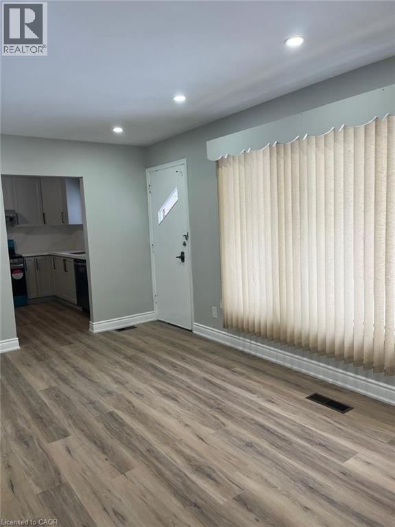Spacious room featuring wood-finish flooring, recessed ceiling lighting, a white entry door, and a large window with vertical blinds - 102 Massey Avenue, Kitchener, ON