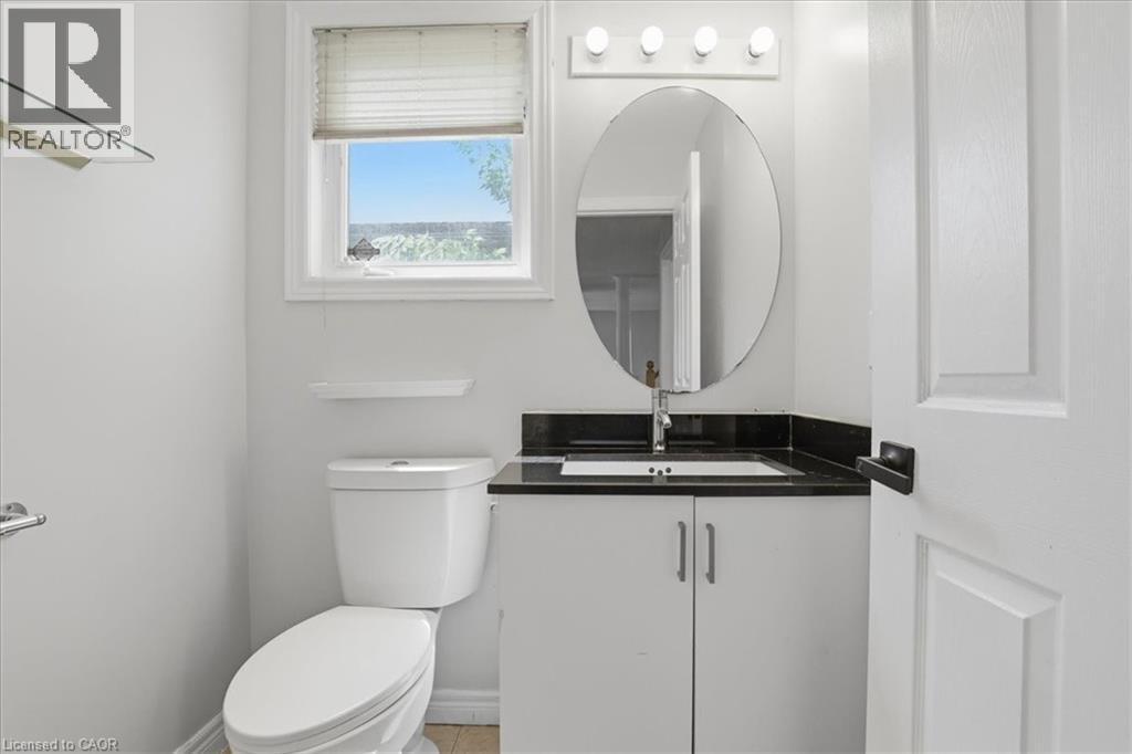 Bright white powder room featuring a window with privacy blind, an oval mirror, a dark countertop vanity with cabinet storage, and a white toilet - 43 Decaro Crescent, Cambridge, ON