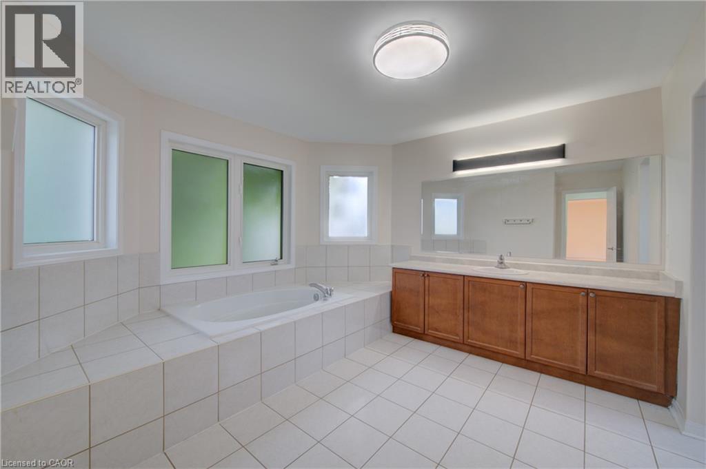Spacious bathroom featuring a built-in soaking tub surrounded by white tile, multiple frosted windows, and a wood-finish vanity with an integrated sink and an expansive mirror - 180 Arthur Fach Drive, Cambridge, ON
