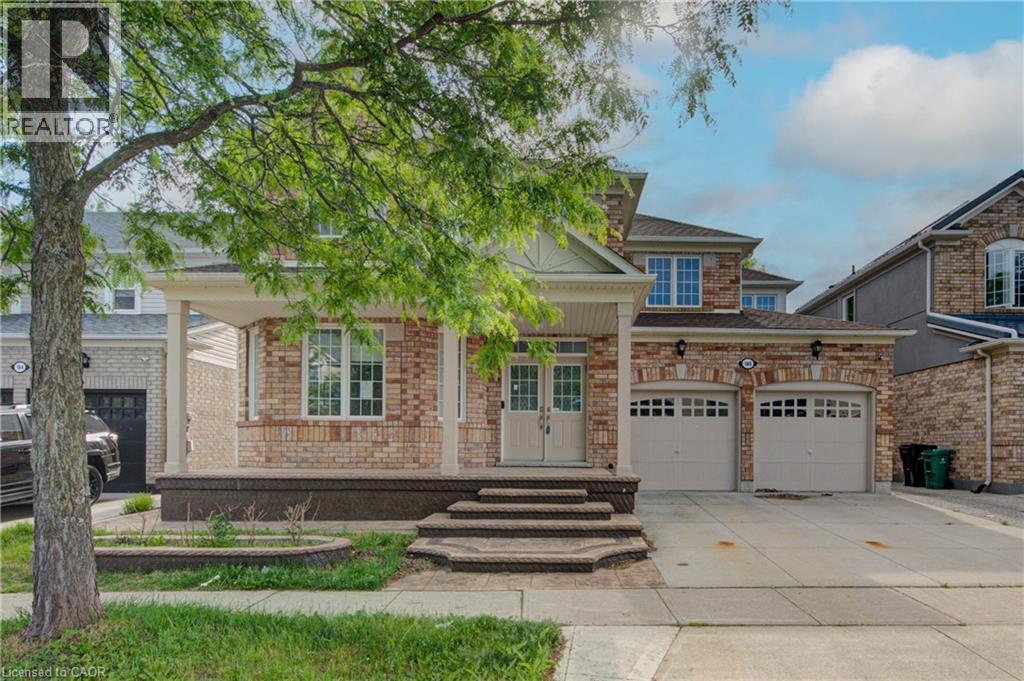 Brick-clad residence featuring a covered front porch, double entry doors, a two-car garage with carriage-style doors, a paved driveway, and mature landscaping - 180 Arthur Fach Drive, Cambridge, ON