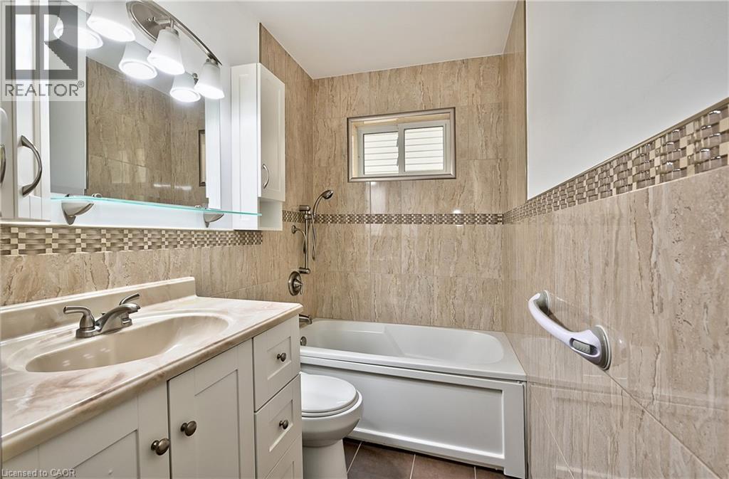 Bathroom featuring a vanity with an integrated sink and light-toned countertop, a full bathtub with a showerhead, beige tiled walls, a decorative mosaic tile accent band, and a window with horizontal - 1476 Barton Street E Unit# 2, Hamilton, ON