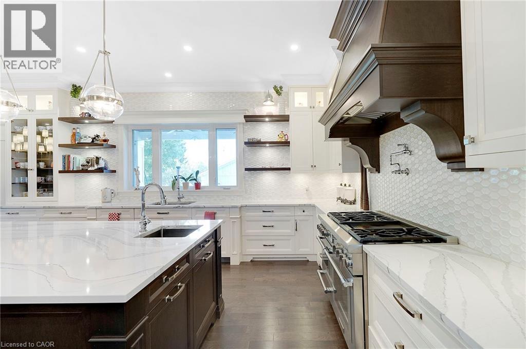 Gourmet kitchen featuring a large island with an undermount sink, white cabinetry, dark wood-finish range hood, white patterned tile backsplash, and stainless steel appliances - 6195 Milburough Line, Burlington, ON - Indoor Photo Showing Kitchen With Upgraded Kitchen