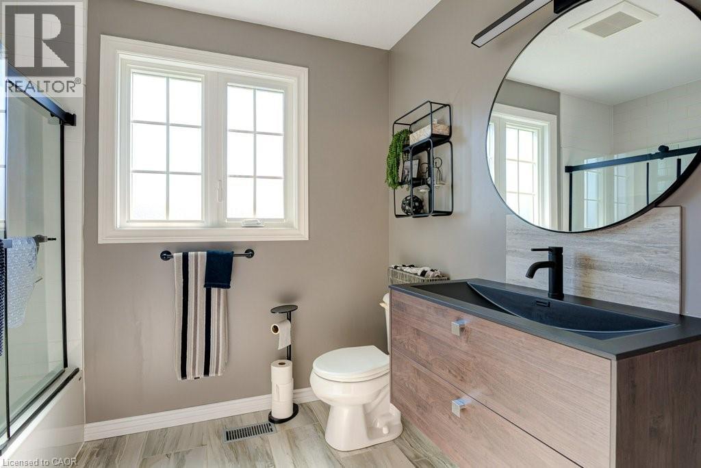 Bathroom featuring a wood-finish vanity with an integrated basin, a large circular mirror, and a window with grid detailing - 61 Ditner Avenue, Baden, ON - Indoor Photo Showing Bathroom