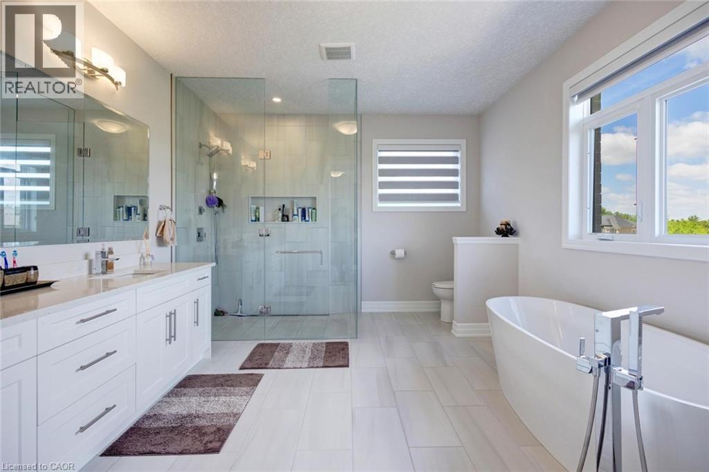 Contemporary bathroom featuring a freestanding tub, glass-enclosed shower, dual vanity with white cabinetry, light-toned tile flooring, and multiple windows - 57 Hollybrook Trail, Kitchener, ON