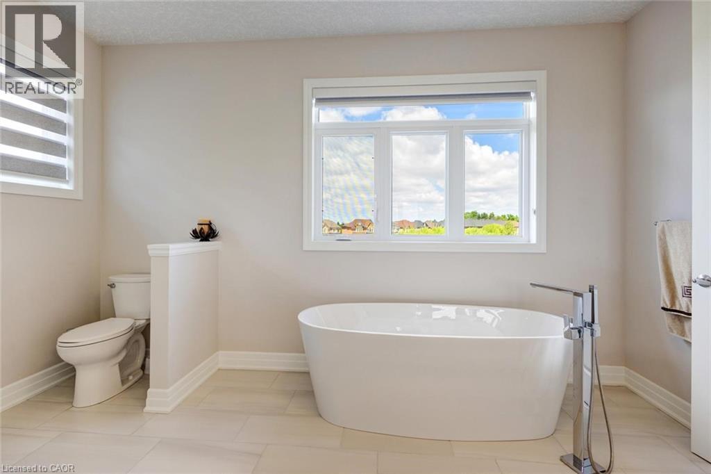 Freestanding soaking tub with a floor-mounted chrome faucet, large window, separate toilet, and light-toned tile flooring - 57 Hollybrook Trail, Kitchener, ON