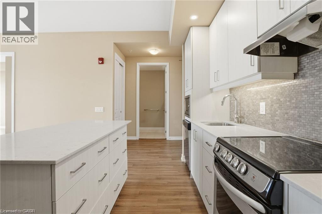 Modern kitchen featuring a white countertop island, white cabinetry, a built-in range, a stainless steel sink, and a gray tile backsplash - 40 Esplanade Lane Unit# 614, Grimsby, ON