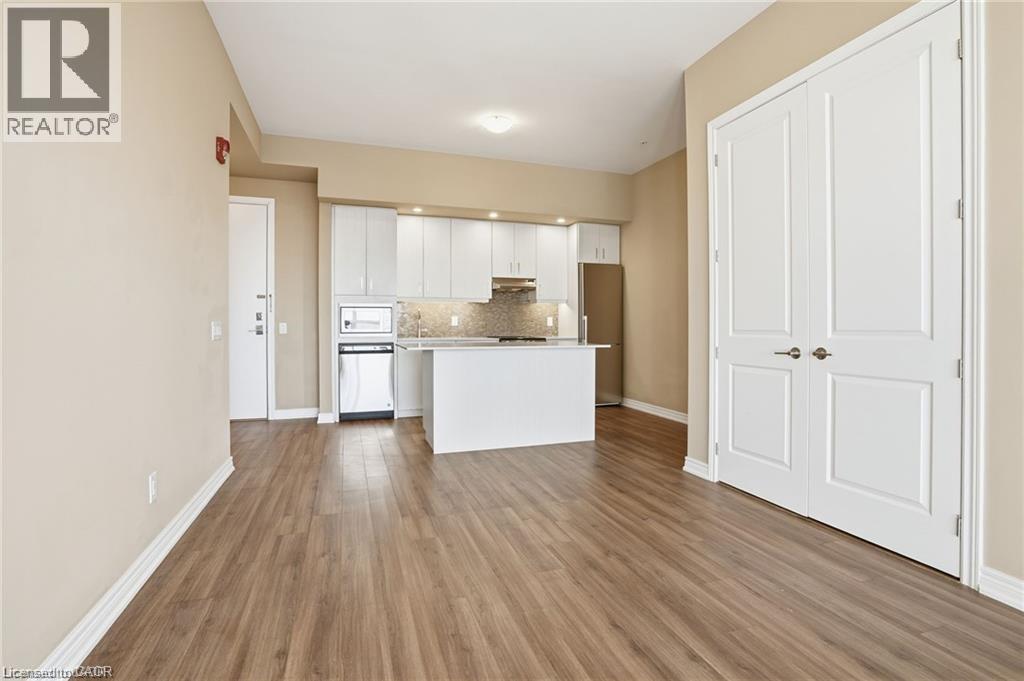 Open-concept living area featuring wood-finish flooring, a contemporary kitchen with white cabinetry and an island, a stainless steel refrigerator, and a built-in microwave - 40 Esplanade Lane Unit# 614, Grimsby, ON