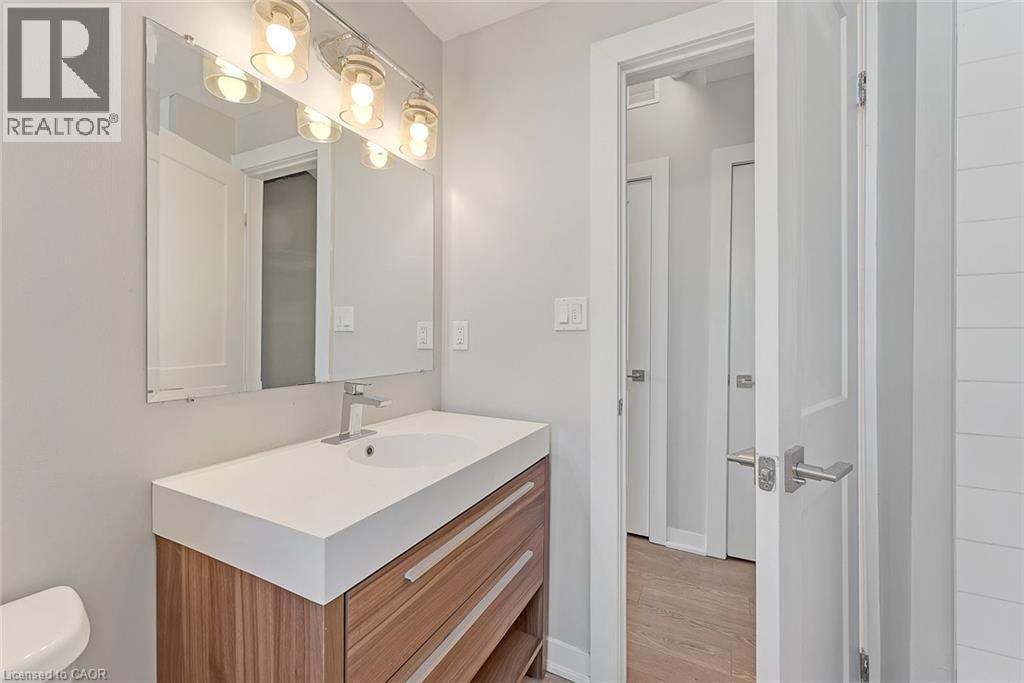 Contemporary bathroom featuring a wood-finish vanity with a white countertop and integrated sink, brushed nickel faucet, large frameless mirror, and a modern four-light fixture - 657 Barton Street E Unit# 302, Hamilton, ON