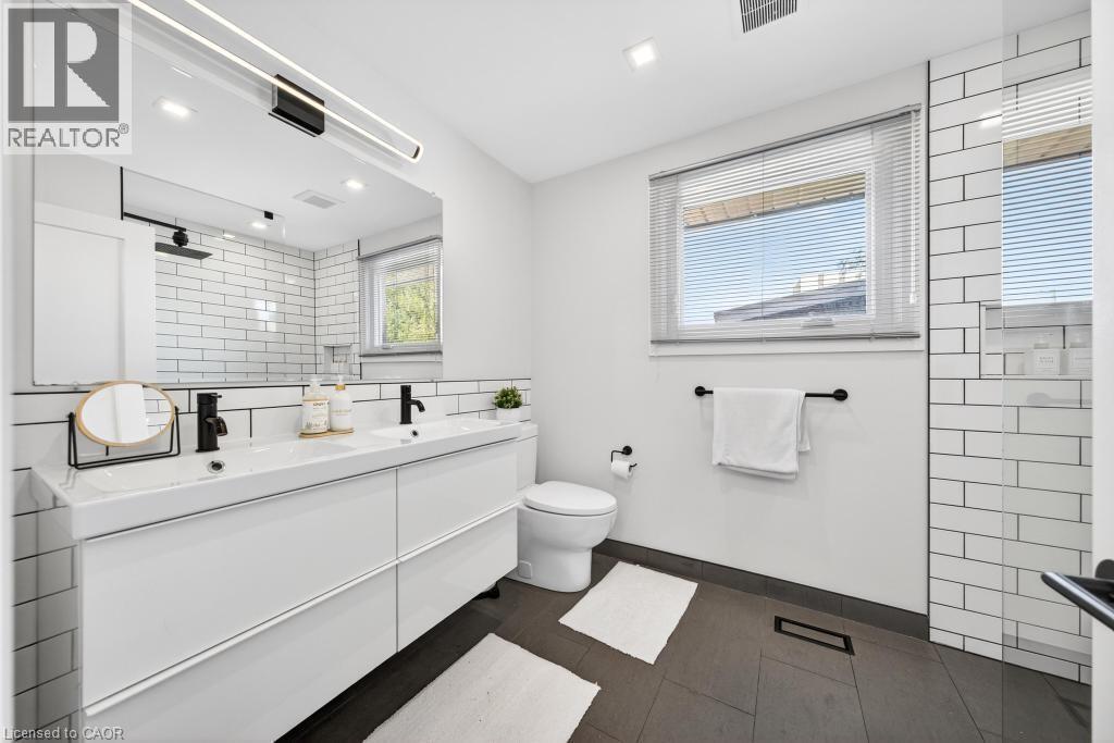 Contemporary bathroom featuring a dual vanity with integrated sinks, matte black fixtures, a large mirror with integrated lighting, a window with horizontal blinds, and subway tile accents - 19 Glen Forest Drive, Hamilton, ON