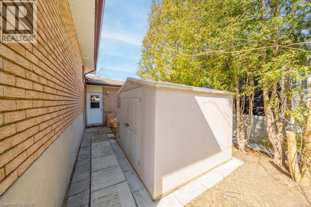 Side yard featuring a brick exterior wall, a paved walkway, a white utility shed, a white entry door with a window, and mature trees - 19 Glen Forest Drive, Hamilton, ON
