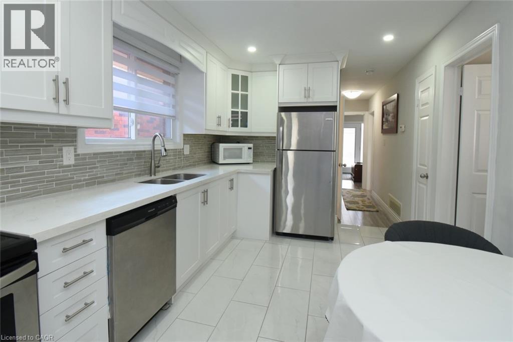 Kitchen featuring white cabinetry, light-toned countertops, a stainless steel double-basin sink, stainless steel appliances, and a gray subway tile backsplash - 185 Rosslyn Avenue N, Hamilton, ON - Indoor Photo Showing Kitchen With Double Sink With Upgraded Kitchen