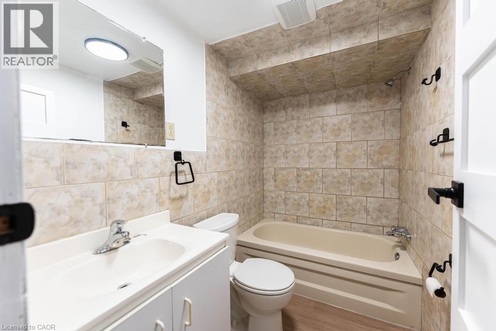 Bathroom featuring a ceramic tile bath surround, a white vanity with integrated sink, and wood-finish flooring - 65 Garden Crescent Unit# Lower, Hamilton, ON - Indoor Photo Showing Bathroom
