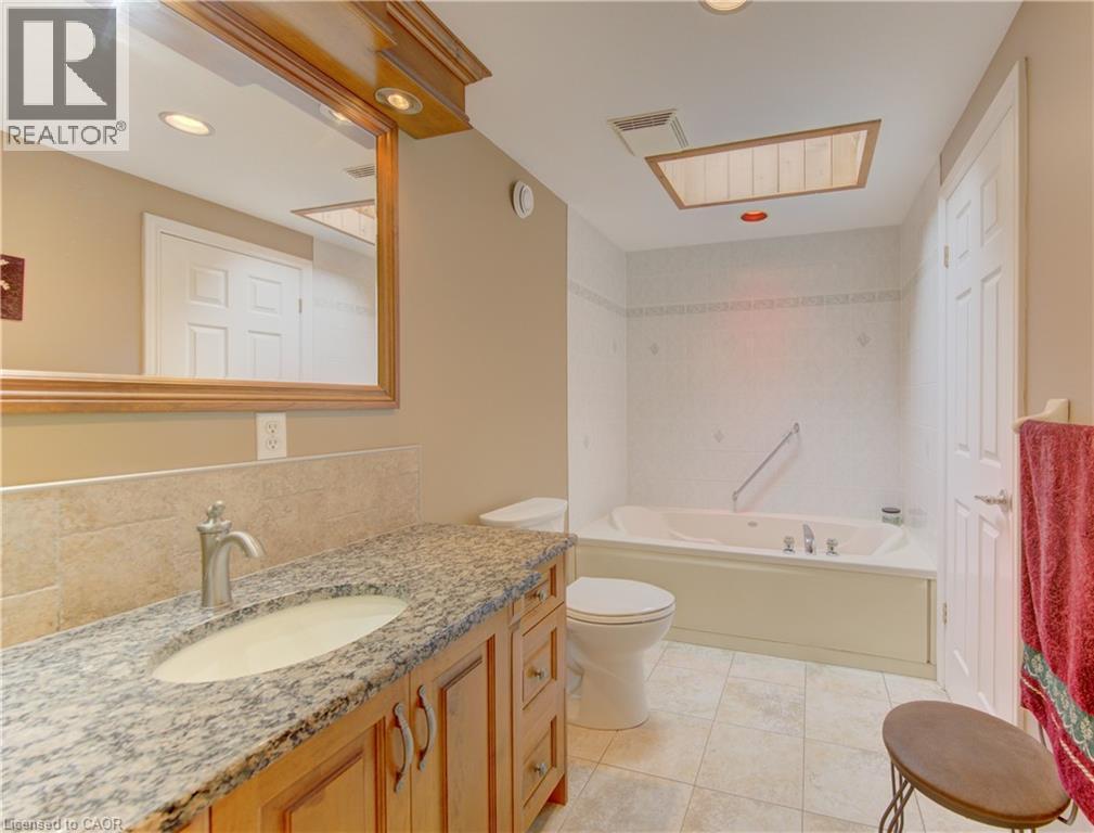 Bathroom featuring a large jetted tub, a vanity with a granite-style countertop, a single basin sink, and a framed mirror with integrated lighting - 599 Coldstream Drive, Waterloo, ON