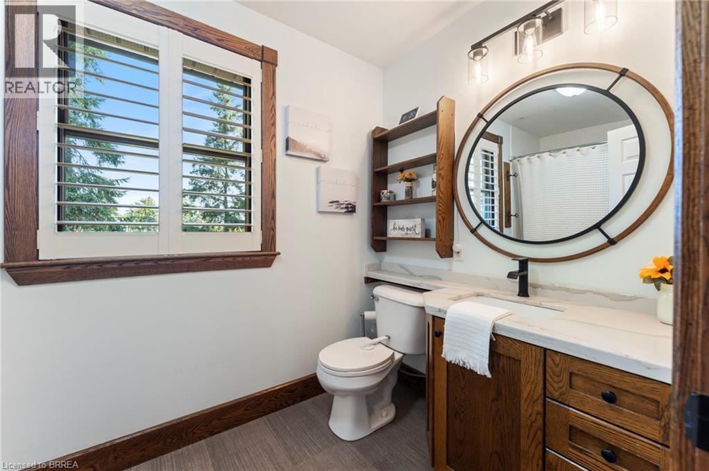Bathroom featuring a wood-finish vanity with a light-colored countertop, a circular framed mirror, and a window with plantation shutters - 182 Governors Road E, Paris, ON