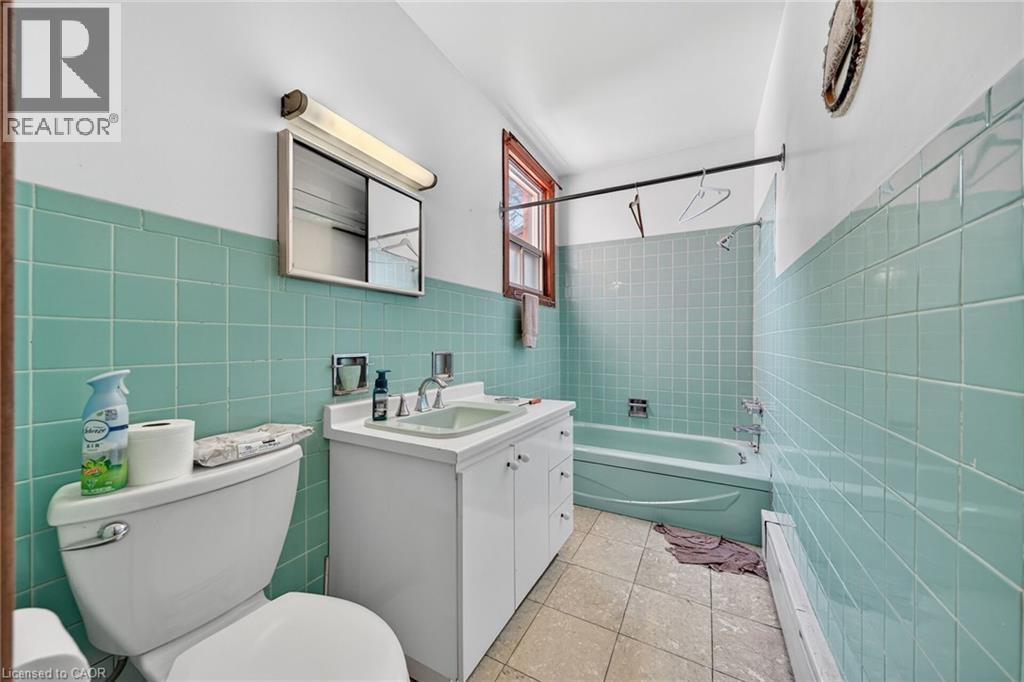 Bathroom featuring teal wall tiling, a white vanity with integrated sink, a white toilet, a built-in bathtub, and a window with wood trim - 16 Mulberry Street, Hamilton, ON
