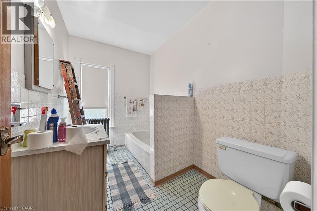 Bathroom featuring a vanity with an integrated sink, a built-in bathtub, a toilet, tiled walls, and a window with a roller shade - 16 Mulberry Street, Hamilton, ON