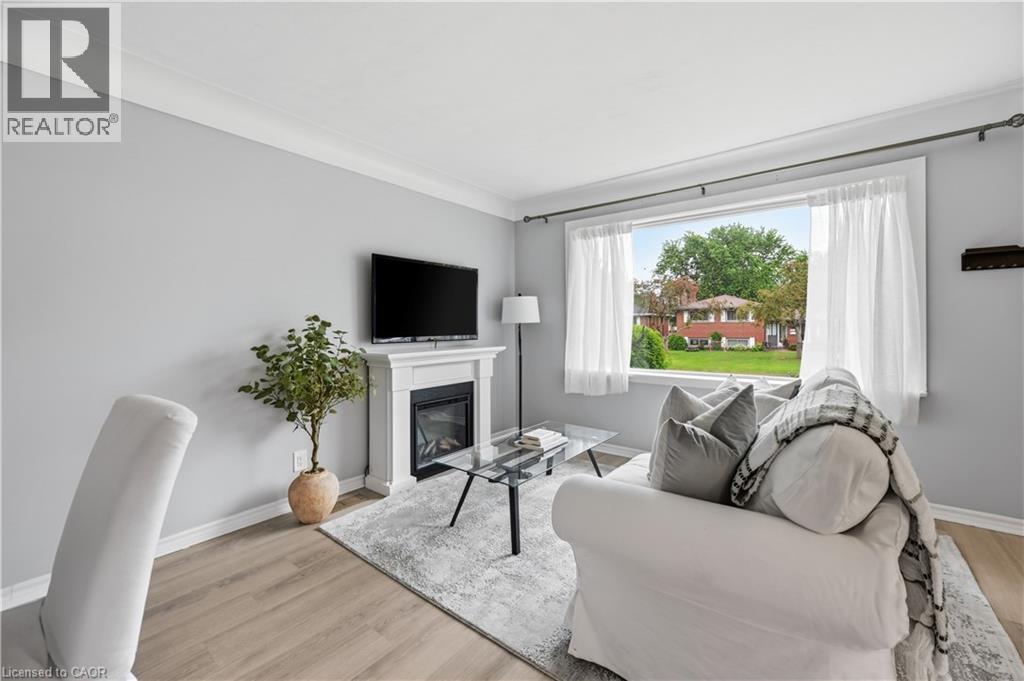 Living area featuring a fireplace with a white mantel, light grey walls, white crown molding, wood-finish flooring, and a large window - 189 Foxbar Road, Burlington, ON