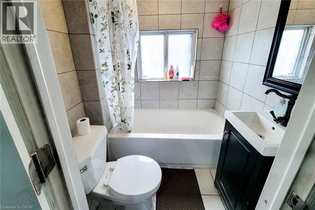 Bathroom featuring a built-in bathtub with tiled surround, a window, a single vanity with dark cabinetry, and a toilet - 463 Wellington Street N, Hamilton, ON - Indoor Photo Showing Bathroom