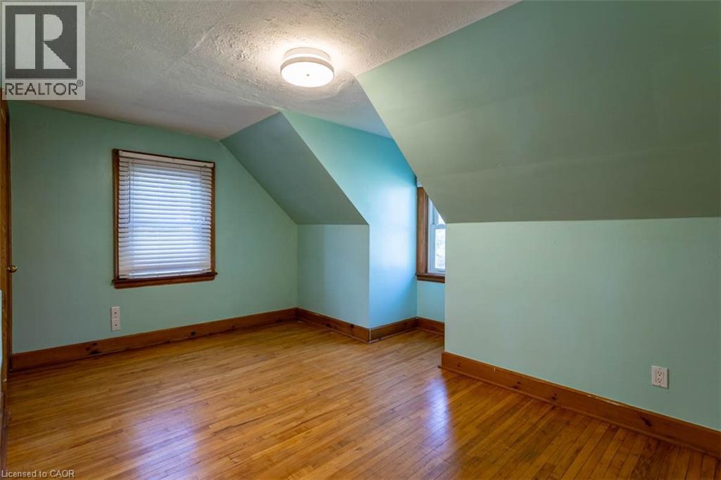 Room featuring wood-finish flooring, two windows with wood trim, and a ceiling-mounted light fixture - 17 Thorndale Street N, Hamilton, ON