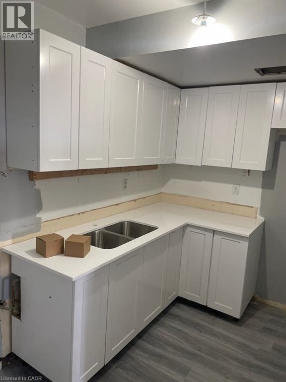 Corner kitchen area featuring white shaker-style cabinetry, a double-basin stainless steel sink, light-toned countertops, and wood-finish flooring - 150 Charlton Avenue E Unit# 708, Hamilton, ON