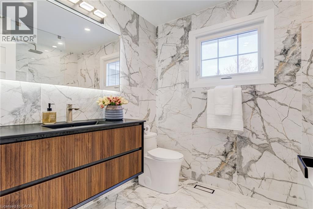 Bathroom featuring large-format marble-look wall and floor tiling, a floating wood-finish vanity with a dark countertop and integrated sink, a large mirror with integrated lighting, and a contemporary - 1320 Monmouth Drive, Burlington, ON