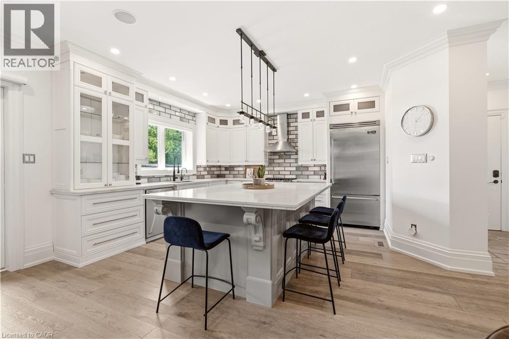 Gourmet kitchen featuring a large central island, light wood-finish flooring, white shaker-style cabinetry with glass accents, stainless steel appliances, and a subway tile backsplash - 336 Newbold Court, Burlington, ON