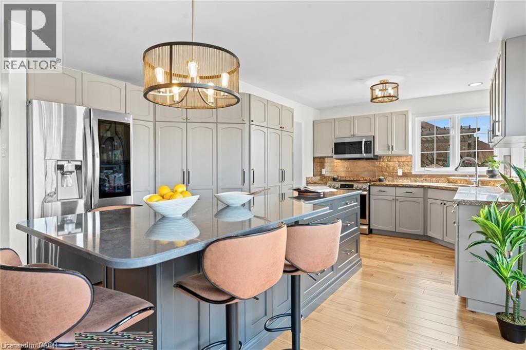 Kitchen featuring a spacious island with seating, stainless steel refrigerator, light gray cabinetry, wood-finish flooring, and a window above the sink - 26 Shadyridge Place, Kitchener, ON