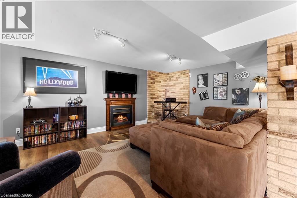 Spacious recreational room featuring wood-finish flooring, a corner fireplace with a detailed mantel, and a multi-tone brick accent wall - 26 Shadyridge Place, Kitchener, ON