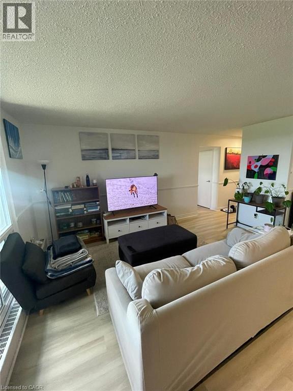 Spacious living area featuring light wood-finish flooring, white wall paint, and a textured ceiling - 225 Harvard Place Unit# 1505, Waterloo, ON